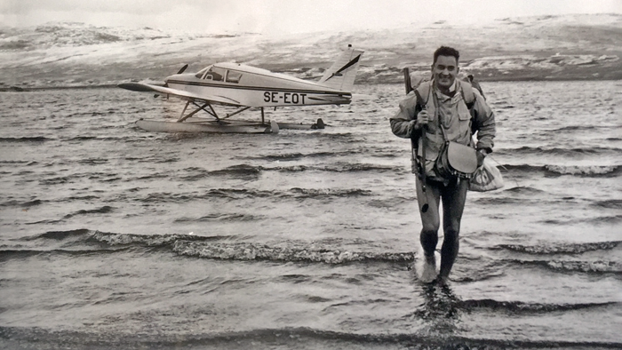 Svartvit bild på ung Göran Gustafsson på en strand, bakom honom i vattnet ett flygplan.
