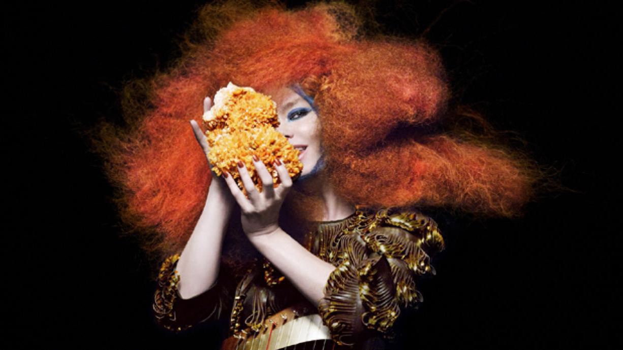 Sångerskan Björk står med orange hår mot en svart bakgrund och håller i korall.