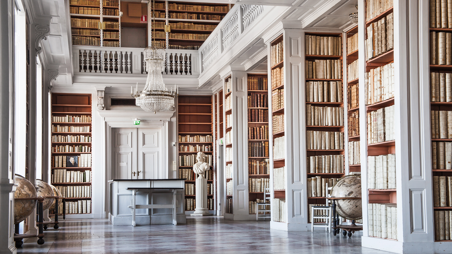 Universitetsbibliotek med vita bokhyllor, byster och historiska jordglober.