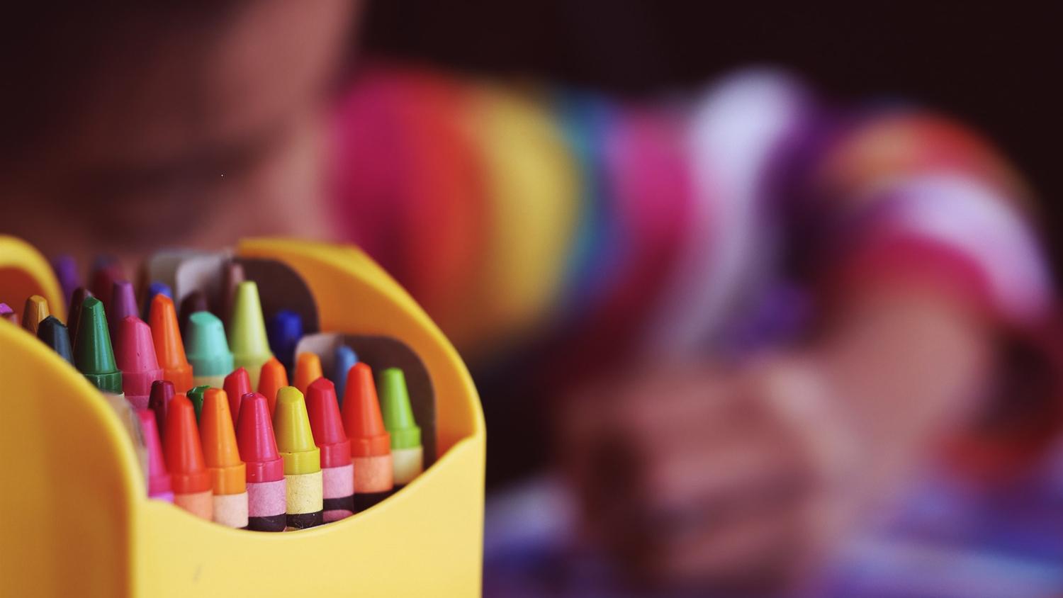 Ask med färgkritor. I bakgrunden syns barn i färgglad tröja som ritar.