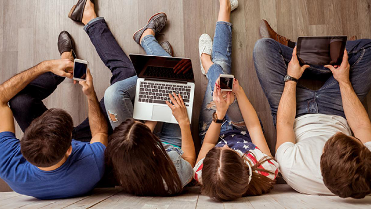 Fyra studenter sitter på golvet lutade mot en vägg med olika mobila enheter i handen.