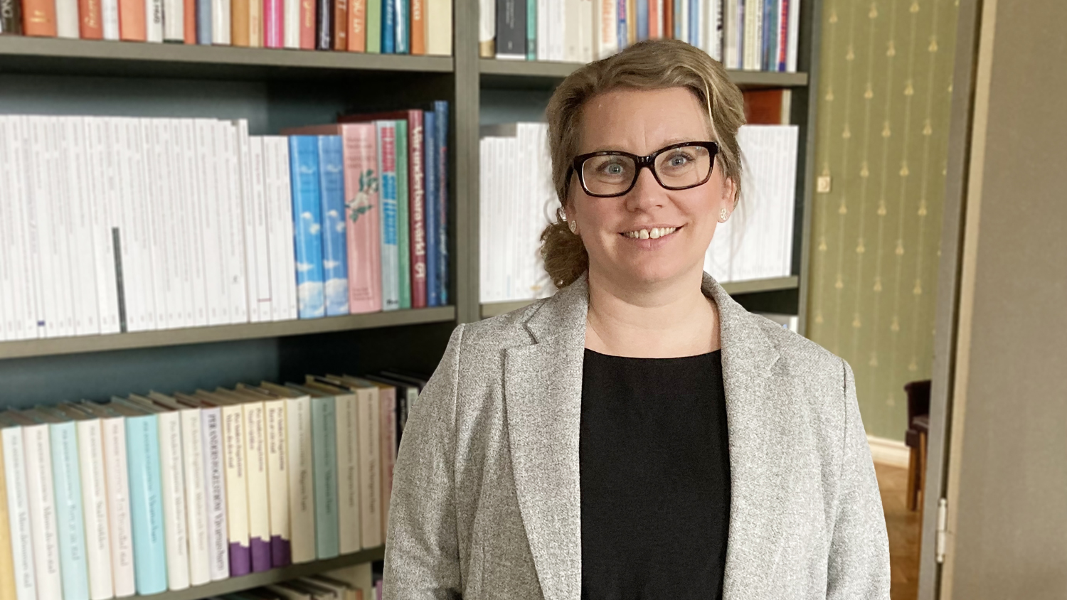 Marlene Malmström har en grå kavaj på sig och står framför en bokhylla.