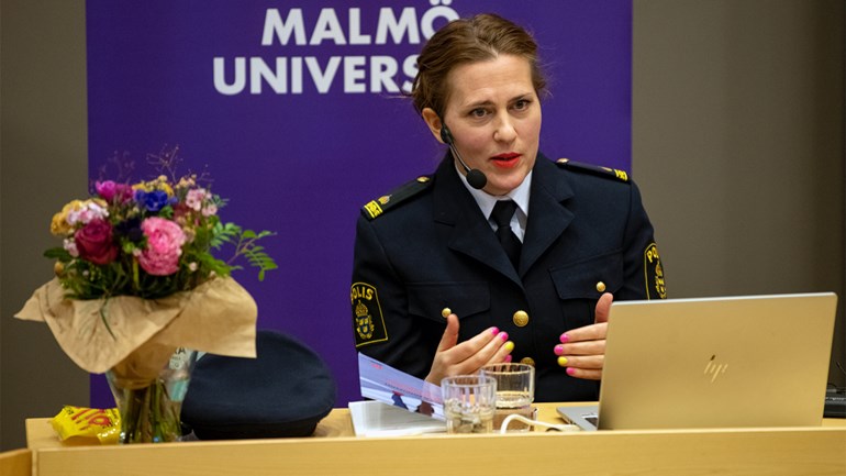 En kvinna i polisuniform sitter vid ett bord framför en dator. Till vänster står en blombukett.
