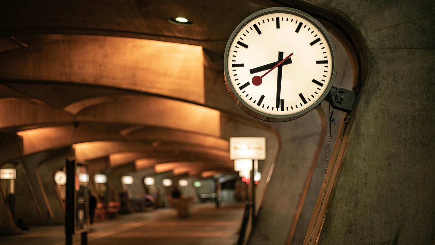 Ett stationsur hänger i förgrunden, brun folktom  lokal som ser ut som en tågstation. 
