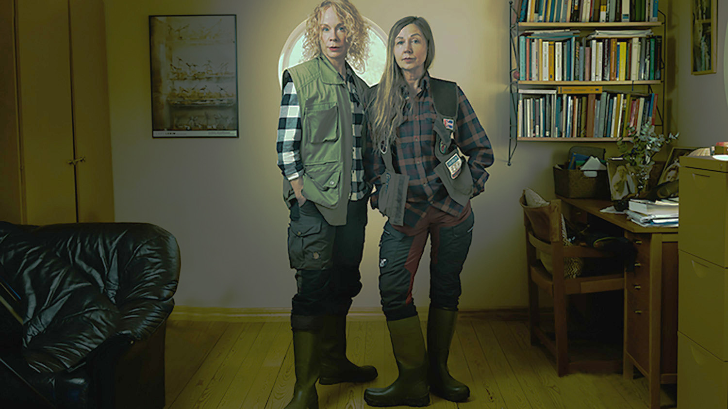 Två kvinnor i jaktväst står bredvid varandra i ett möblerat rum.