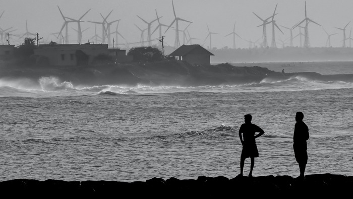 Vy över kustremsa med siluett av två personer i förgrunden och vindkraftverk i bakgrunden. 
