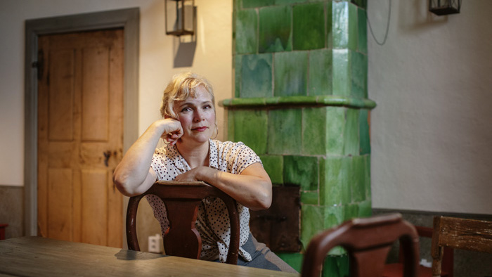 Närbild på Rebecka Lennartsson inomhus där hon sitter på en stol framför en grön kakelugn. 