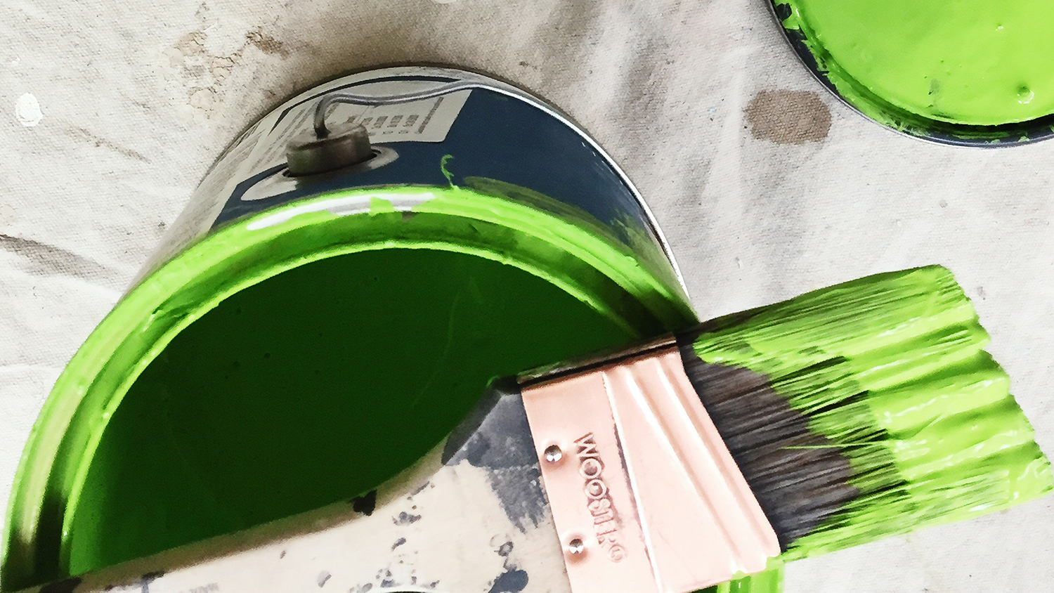 Målarburk med grön färg, ovanpå burken en pensel med färg på, i högra hörnet syns locket med grön färg på. 
