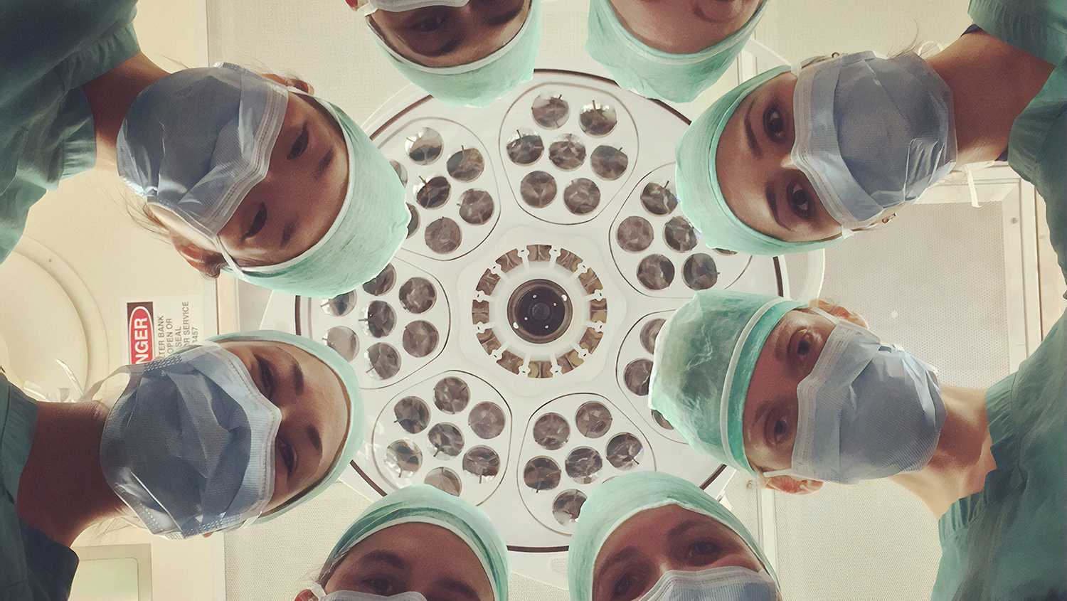 8 personer med kirurgrockar och munskydd böjer sig fram för att titta ner på en patient utanför bild. Ovanför deras huvuden syns taket.