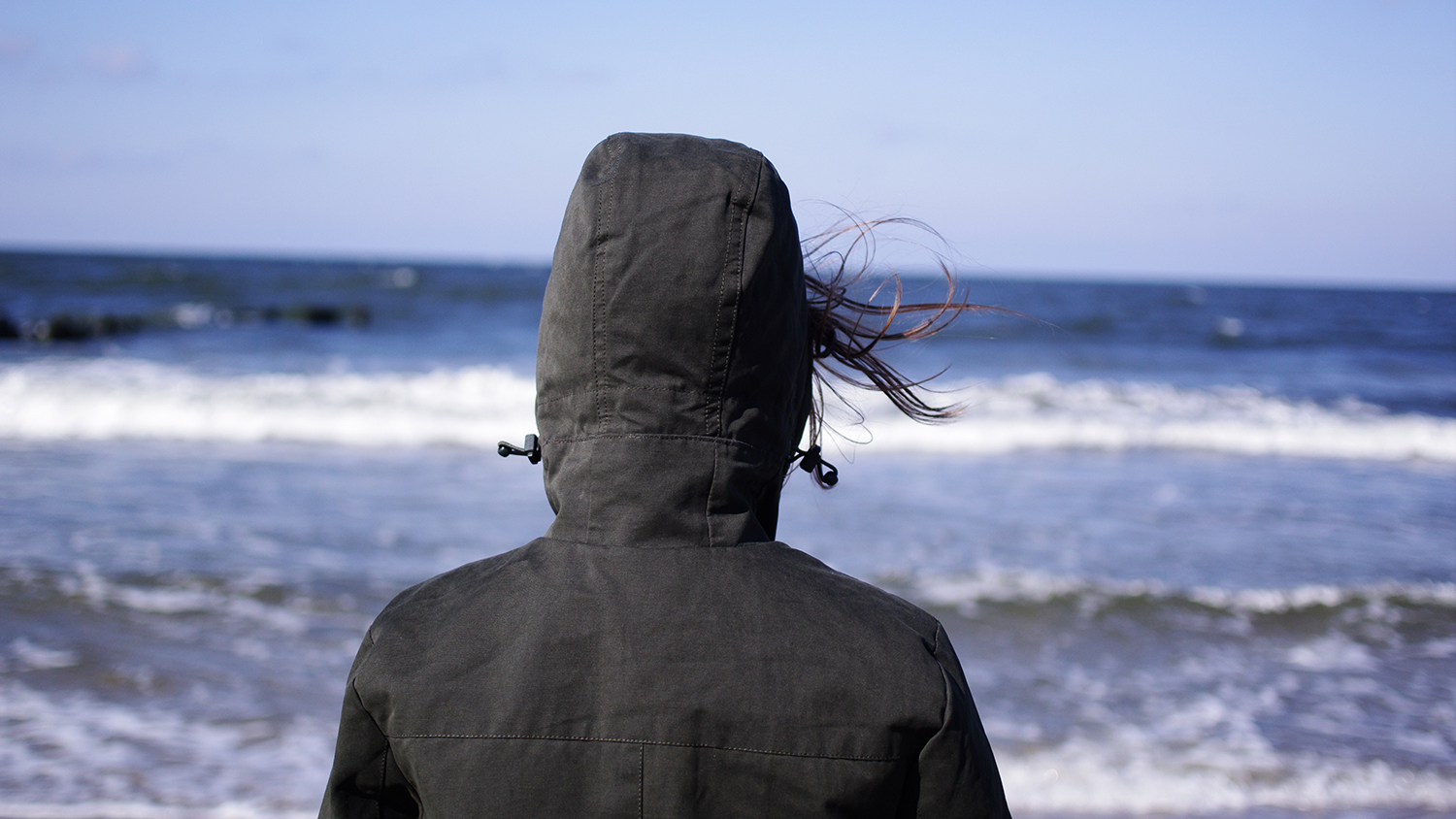 Person fotograferad bakifrån står vid stormande hav iklädd  jacka med kapuschong.