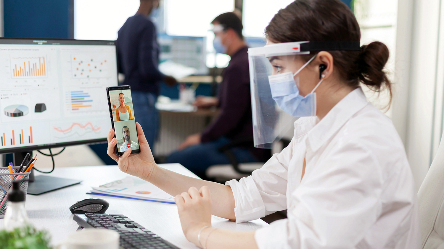 Kvinna med munskydd sitter framför datorn och har videosamtal över mobilen.