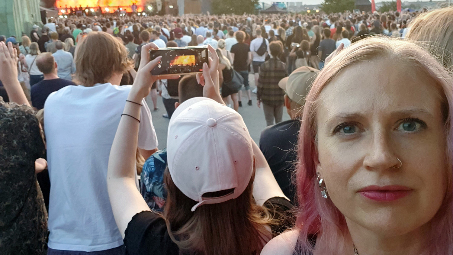 Kvinna med rosafärgat hår tittar in i kameran. I bakgrunden en utomhusscen där det pågår en konsert.