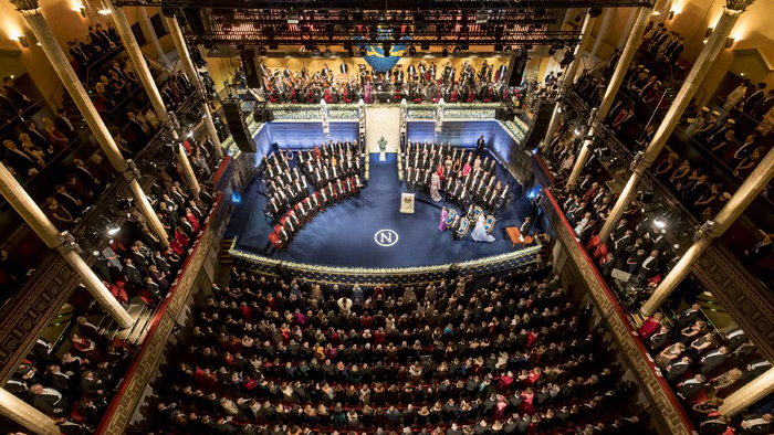Högtidsklädda
Nobelpristagare sitter i halvcirkel på scen, framför en fulltalig publik