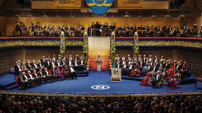 En rad nobelpristagare sitter på en scen med en blå matta.