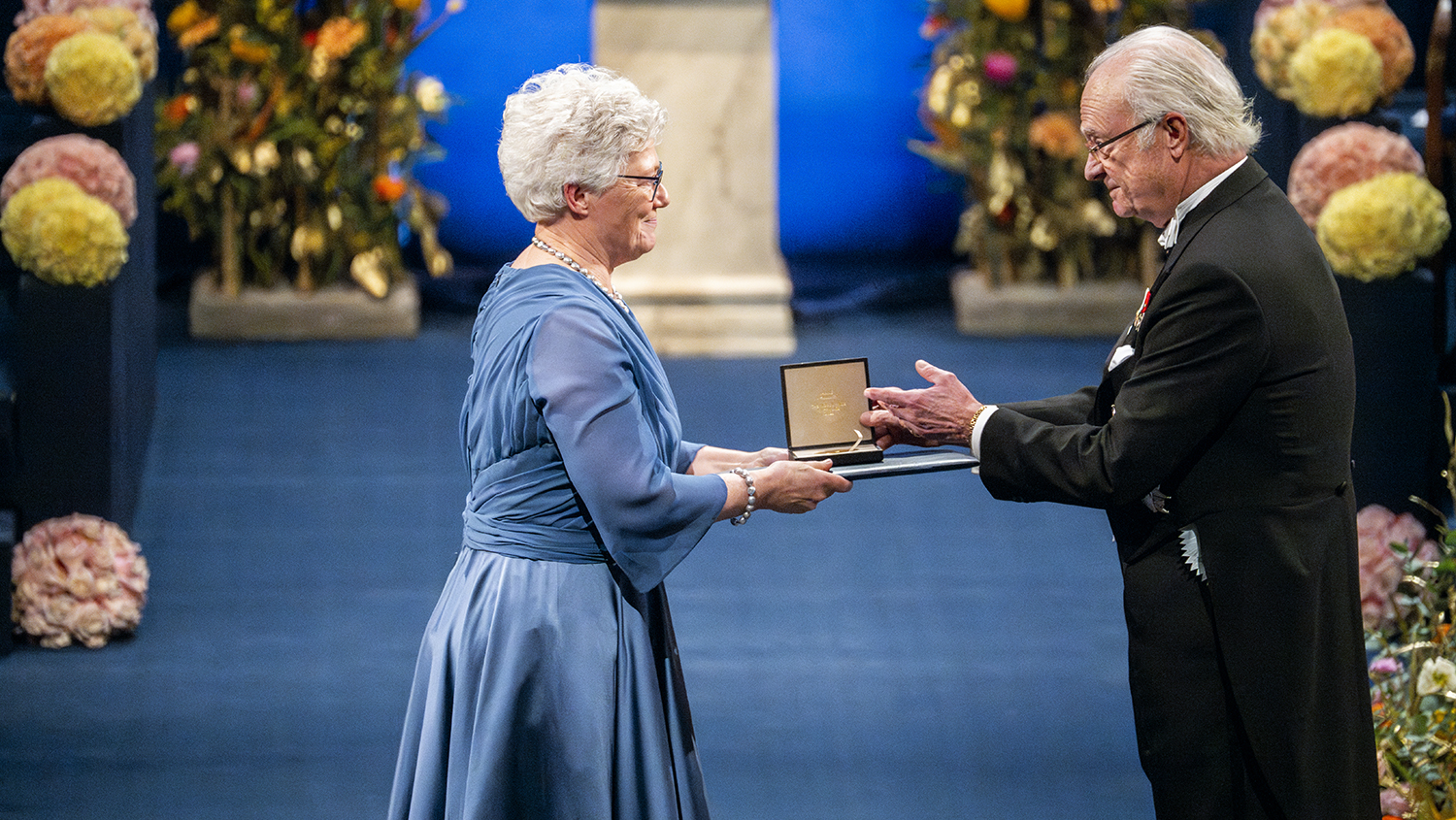 Ann L'Huiller, klädd i blå klänning, tar emot Nobelpriset av kungen. Blå interiör med stora blommor.